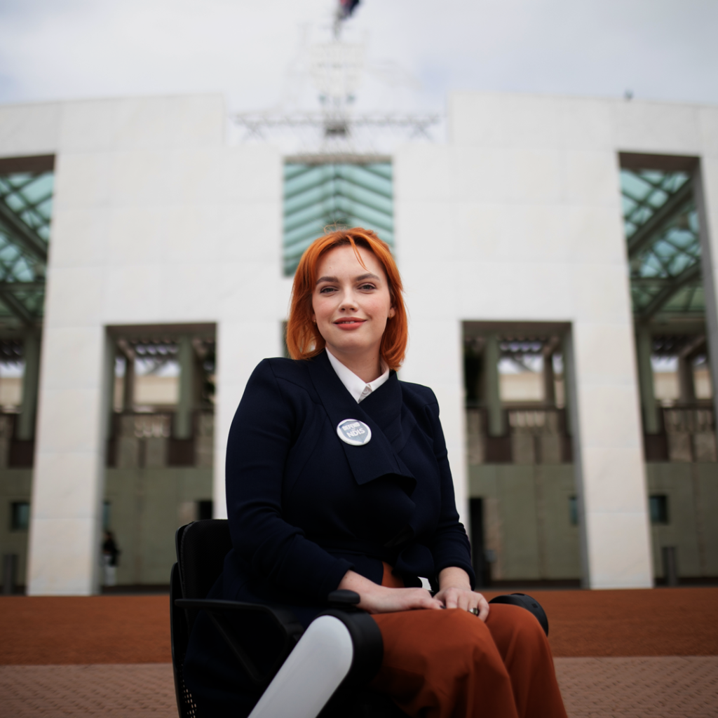 Elly Desmarchelier is pictured in front of the Parliament House in Canberra. Elly is smiling. She is in a wheelchair, and an NDIS badge is pinned to her blazer.