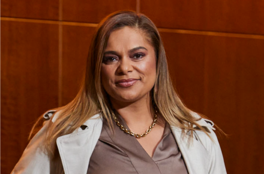 Mundanara Bayles looking down the camera lens. She is wearing a brown shirt, a white jacket and a gold necklace.